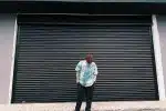 A Man in in Blue Shirt and Black Pants Standing Near Steel Rolling Shutter while Looking Down