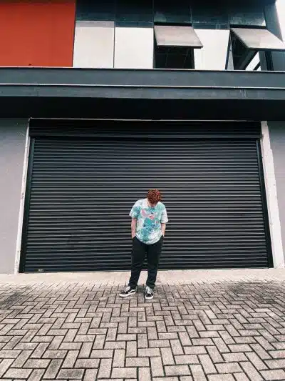 A Man in in Blue Shirt and Black Pants Standing Near Steel Rolling Shutter while Looking Down