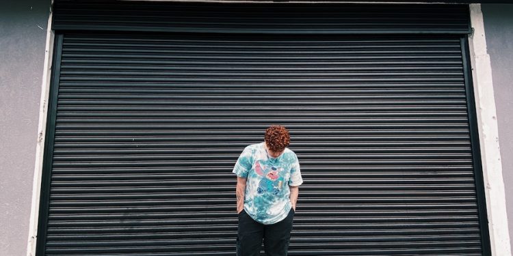 A Man in in Blue Shirt and Black Pants Standing Near Steel Rolling Shutter while Looking Down