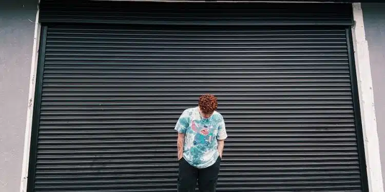 A Man in in Blue Shirt and Black Pants Standing Near Steel Rolling Shutter while Looking Down